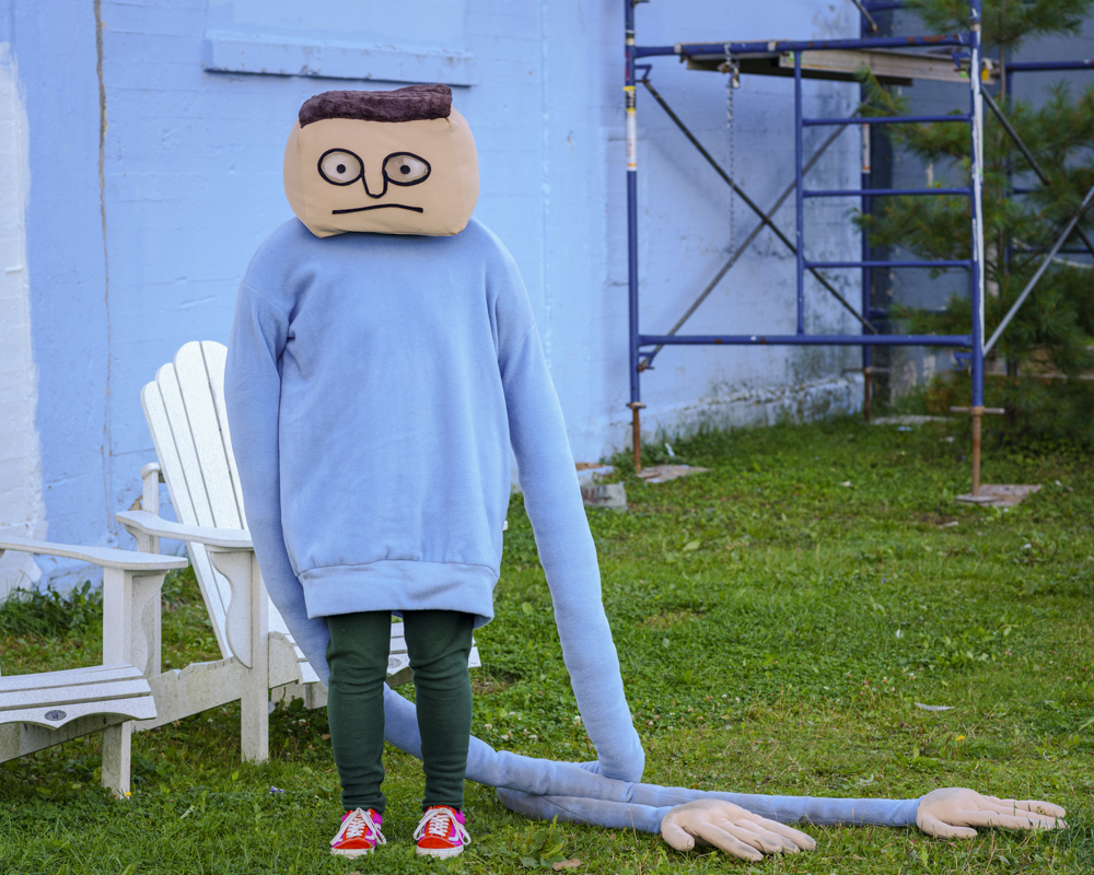 A mascot-like figure with a square head and blue sweater with very long sleeves stands in front of a blue building