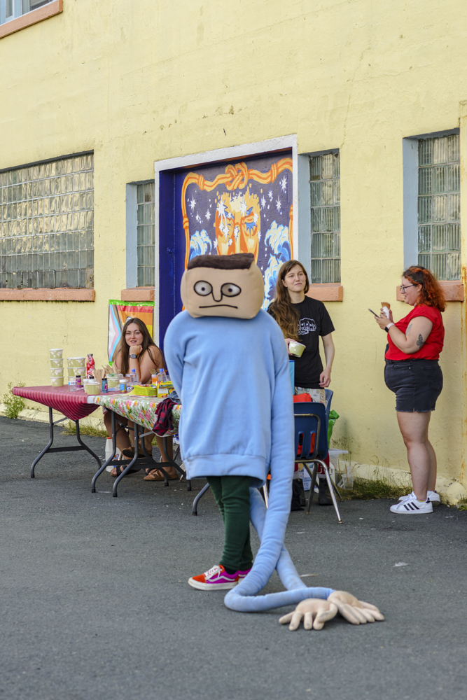 A mascot-like figure with a square head and blue sweater with very long sleeves stands in front of a building