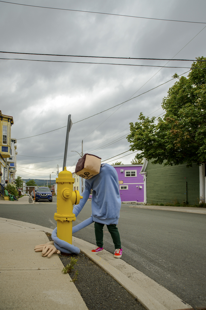 A mascot-like figure with a square head and blue sweater with very long sleeves walking on the sidewalk, tangled in a yellow fire hydrant