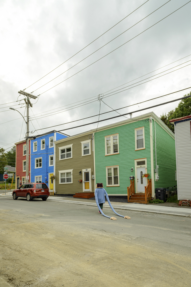 A mascot-like figure with a square head and blue sweater with very long sleeves walking on the sidewalk, behind are colourful houses