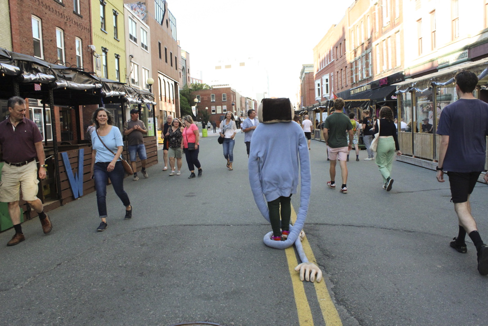 A mascot-like figure with a square head and blue sweater with very long sleeves walking on a downtown road crowded with people