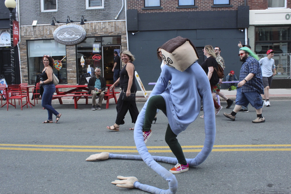 A mascot-like figure with a square head and blue sweater with very long sleeves walking on a road, trying to kick its own arms