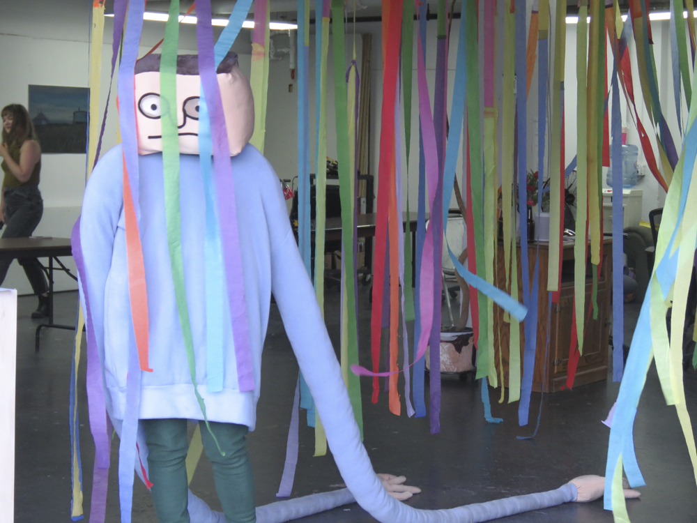 A mascot-like figure with a square head and blue sweater with very long sleeves stands in colourful streamers
